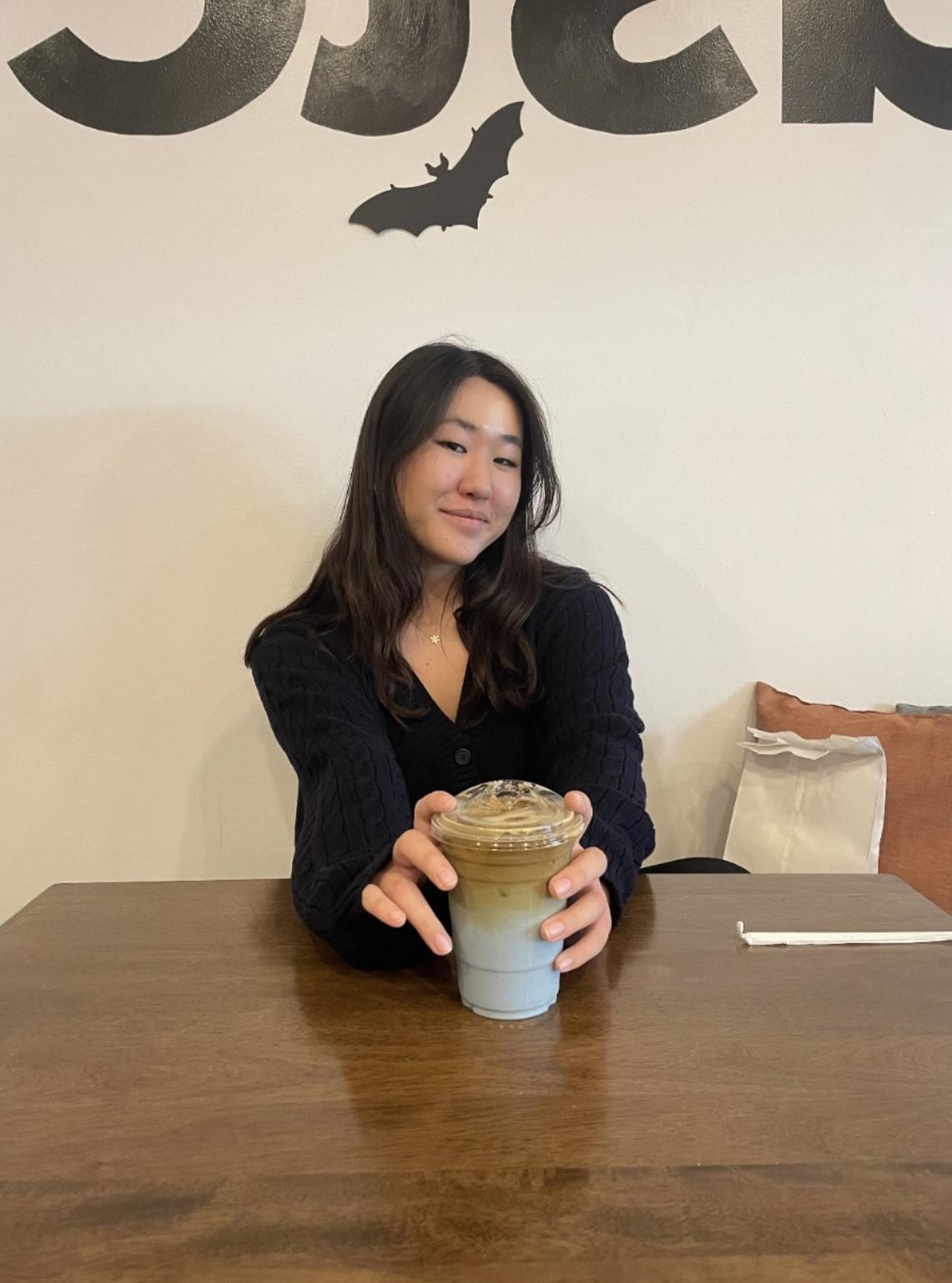 Asian woman wearing black shirt sitting at a table smiling with an iced coffee