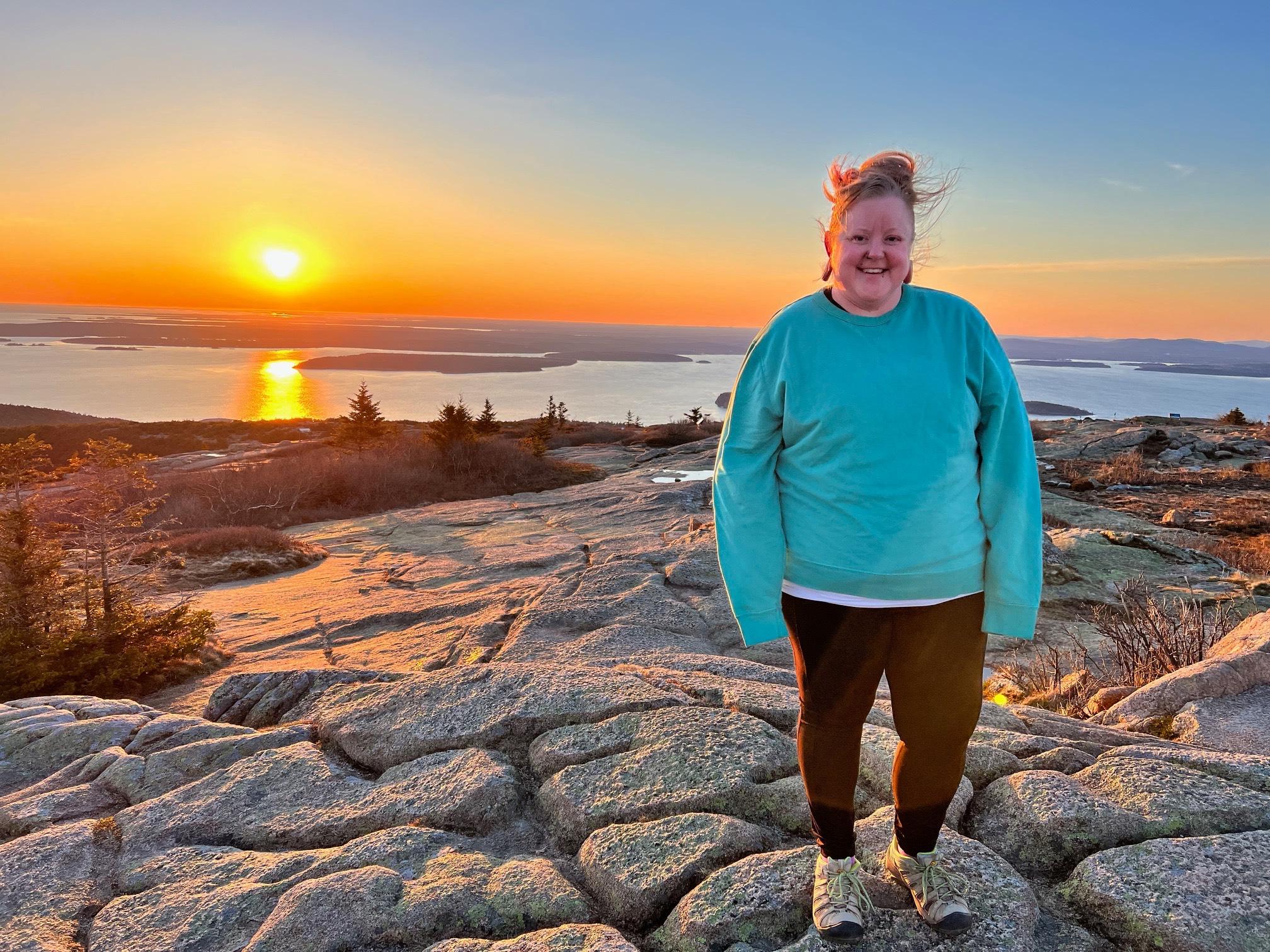 A white woman stands on top of a mountain at sunrise.